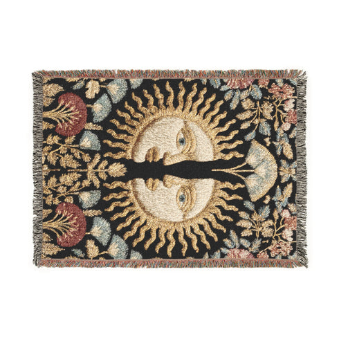 Jacquard-woven cotton blanket featuring twin celestial suns—two serene faces meeting at center—with radiant golden rays on a charcoal ground, bordered by folkloric botanicals and fan blossoms in sage, sky, and garnet. Crafted from 100% soft cotton yarn; the artwork is woven, not printed. Reversible with a color-negative back and finished with a protective 2-inch multicolor fringe. Sizes: 52×37, 50×60, 60×80 for wall, sofa, or queen-bed layering.